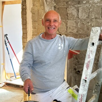 Ein Handwerker lächelt freundlich, während er mit einer Kelle in einer Baustelle steht. Er hält einen Eimer und ist in Arbeitskleidung gekleidet. Im Hintergrund sind unvollendete Wände zu sehen, was auf Renovierungsarbeiten hinweist.