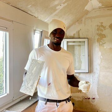 Eine Person mit einem weißen T-Shirt und einer Mütze hält einen Spachtel in der Hand. Der Raum hat unvollendete Wände und Fenster. Diese Person scheint an einem Renovierungsprojekt zu arbeiten.