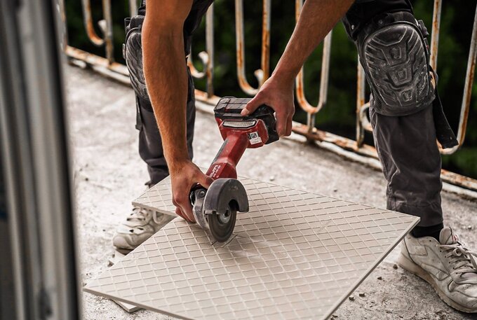 Ein Handwerker schneidet mit einer elektrischen Trennmaschine eine quadratische Fliese mit Struktur. Er hält die Maschine in der einen Hand und die Fliese in der anderen Hand. Der Hintergrund zeigt eine unebene Oberfläche, auf der er arbeitet.