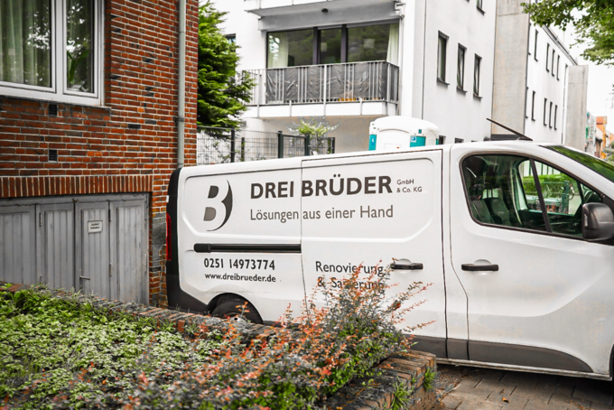 Ein Lieferwagen der Firma "Drei Brüder GmbH & Co. KG" steht an einer Straße. Auf der Seite des Fahrzeugs sind der Firmenname und der Slogan "Lösungen aus einer Hand" zu sehen. Kontaktinformationen und Dienstleistungen wie Renovierung und Sanierung sind ebenfalls aufgeführt.