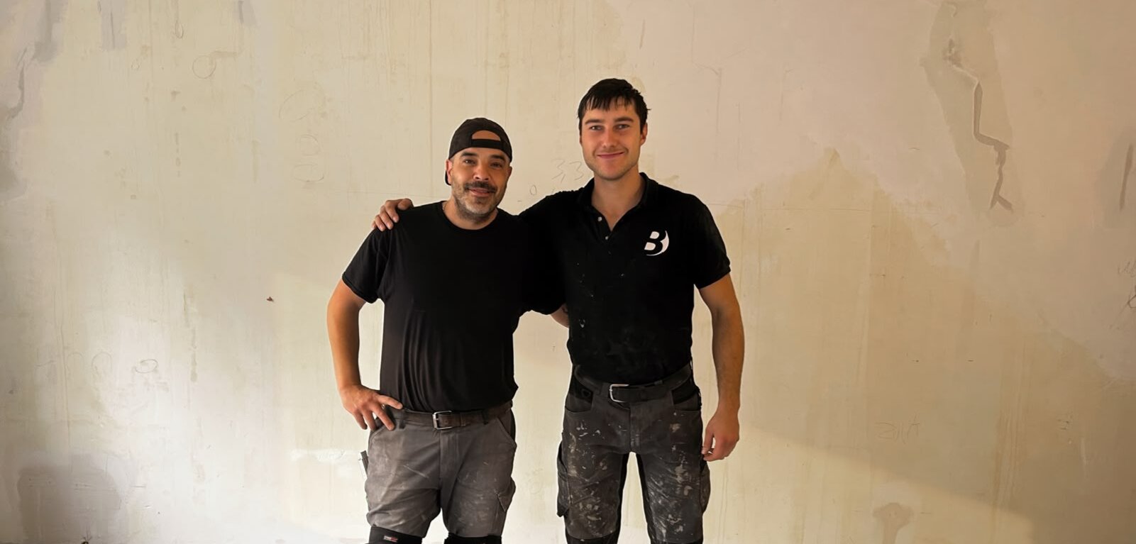 Zwei Handwerker stehen nebeneinander in einem Raum mit unvollendeten Wänden. Beide tragen Arbeitskleidung, einschließlich Kniepolster. Einer hat die Hand auf der Schulter des anderen. Der Boden ist mit Fliesen ausgelegt. Die Umgebung zeigt Bauarbeiten im Gange.