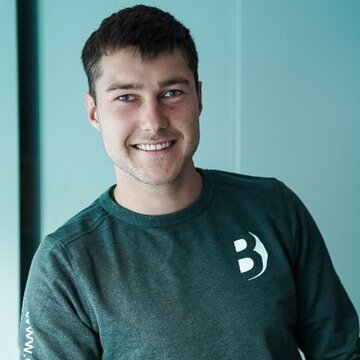 Ein junger Mann lächelt, während er an einem Tisch lehnt. Er trägt ein graues Sweatshirt mit einem Logo. Im Vordergrund sind einige gestapelte Objekte zu sehen, deren Details nicht erkennbar sind. Der Hintergrund ist hell und minimalistisch.