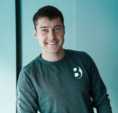 Ein junger Mann lächelt, während er an einem Tisch lehnt. Er trägt ein graues Sweatshirt mit einem Logo. Im Vordergrund sind einige gestapelte Objekte zu sehen, deren Details nicht erkennbar sind. Der Hintergrund ist hell und minimalistisch.