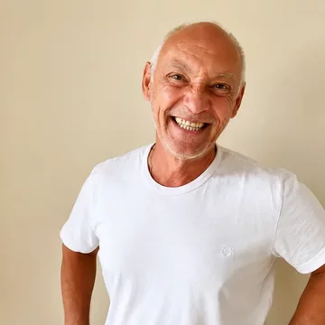 Ein älterer Mann lächelt freundlich in die Kamera. Er trägt ein weißes T-Shirt und hat kurze, graue Haare. Der Hintergrund ist einfarbig beige, was den Fokus auf sein fröhliches Gesicht legt.