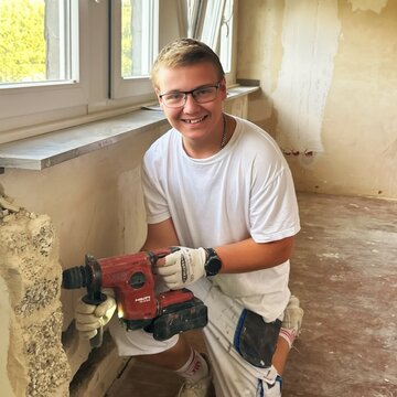 Ein lächelnder junger Mann sitzt auf dem Boden eines renovierungsbedürftigen Raums und hält ein Elektrowerkzeug. Er trägt ein weißes T-Shirt und Handschuhe. Im Hintergrund sind Fenster und unvollendete Wände sichtbar.