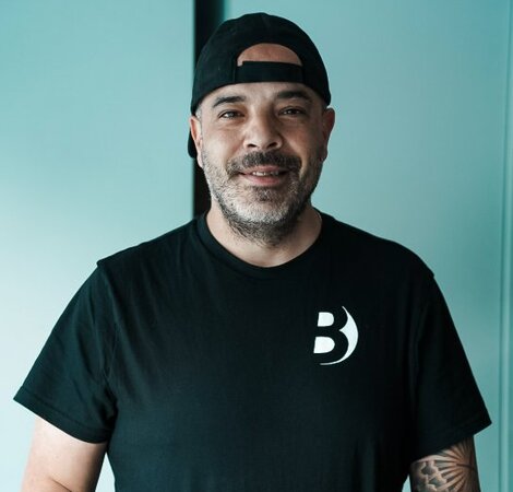 Ein Mann mit einem kurzen Bart und einem schwarzen T-Shirt steht an einem Tisch mit bunten Unterlagen. Er trägt eine Baseballcap und lächelt freundlich in die Kamera. Der Hintergrund ist hell und minimalistisch.