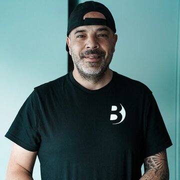 Ein Mann mit einem kurzen Bart und einem schwarzen T-Shirt steht an einem Tisch mit bunten Unterlagen. Er trägt eine Baseballcap und lächelt freundlich in die Kamera. Der Hintergrund ist hell und minimalistisch.